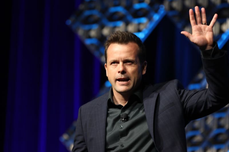 A speaker in a dark jacket presenting on stage with one hand raised.