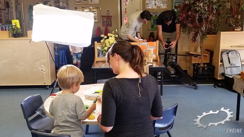 A crew preparing a camera and light while an adult and child work at a table in an educational setting.