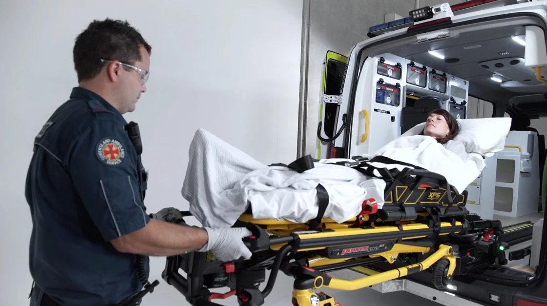 Paramedic demonstrating an ambulance stretcher beside a patient during a training video