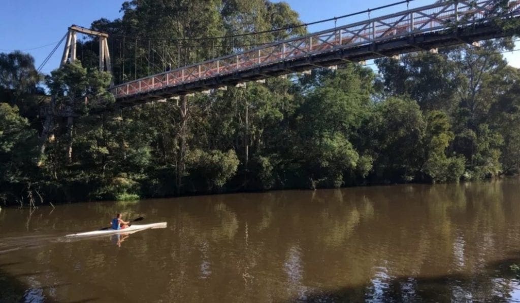 A suspension footbridge over a river, with a paddler below and trees along the banks.