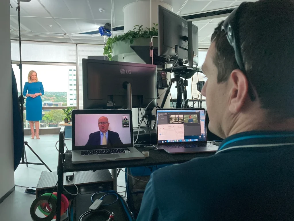 Technician monitors livestream laptops while presenter stands in office studio
