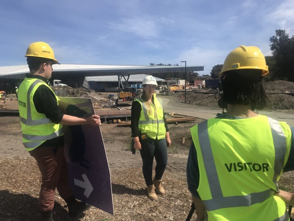 A contributor in high-visibility clothing and a hard hat being filmed outdoors, with a crew member holding a reflector.
