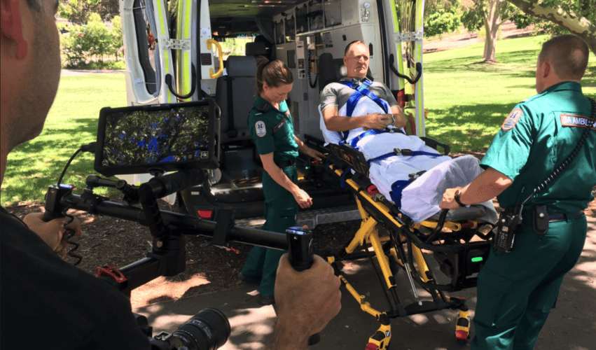 Camera crew filming ambulance personnel demonstrating a Stryker stretcher beside an ambulance