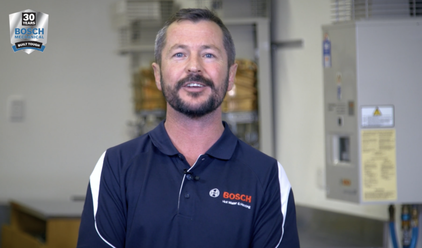 Bosch Hot Water trainer presenting a technical training video beside Bosch continuous flow hot water systems during a commissioning demonstration.