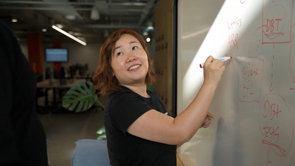 A woman writing on a whiteboard while looking towards a colleague in an office.
