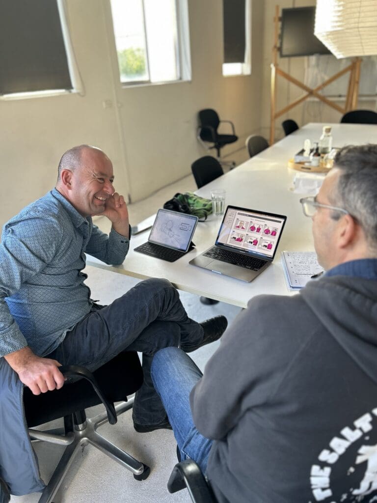 Two people reviewing visual plans on a laptop at a table, with a tablet and printed notes beside it.