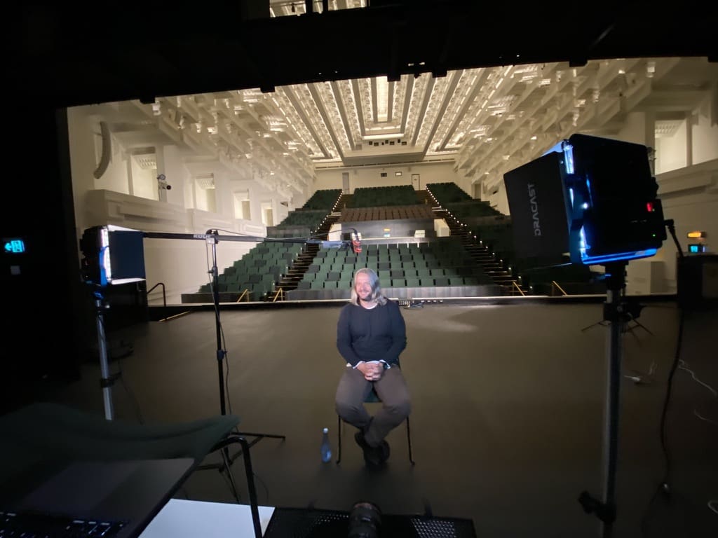 A seated interviewee between lights and a boom microphone in a theatre auditorium.