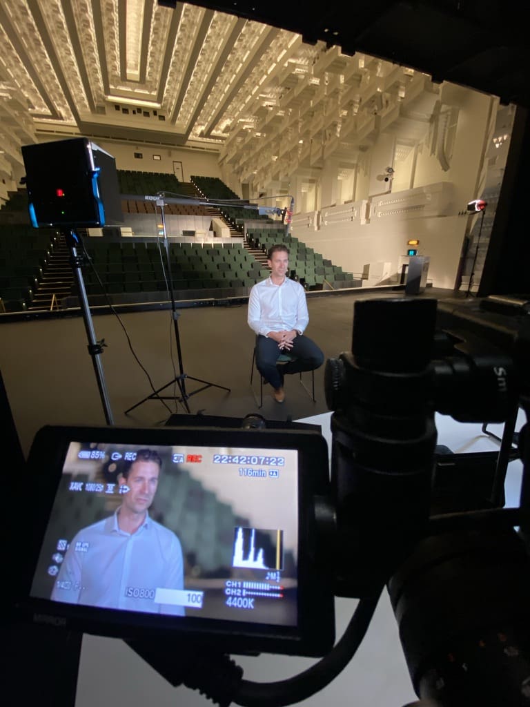 A seated interview subject, camera monitor and lighting in an auditorium.